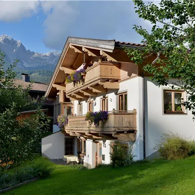 A traditional alpine house with balconies and flowers. In the background, majestic mountains and a blue sky can be seen.