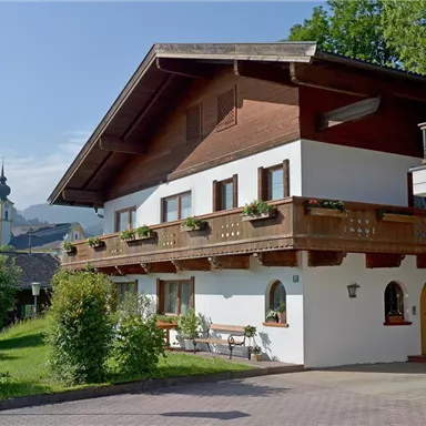 A traditional alpine house with a wooden balustrade and flower boxes. In the background, green hills and a church tower can be seen.