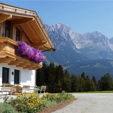 Ein schönes alpenländisches Haus mit einem Balkon, der mit Blumen geschmückt ist. Im Hintergrund sind majestätische Berge und eine klare, blaue Himmel zu sehen.
