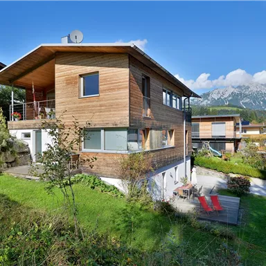 A modern wooden house in a green setting with a view of the mountains. The garden is well-maintained and features a terrace for relaxation.