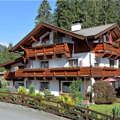 Ein charmantes Holzhaus im alpinen Stil, umgeben von Bäumen und schöner Vegetation. Die Balkone sind mit Blumen dekoriert und das Gebäude strahlt eine einladende Atmosphäre aus.