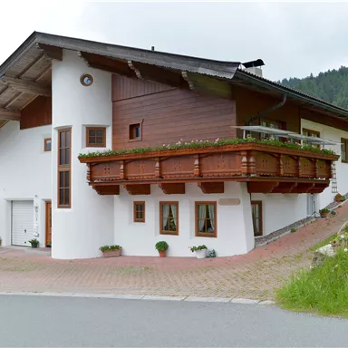 A modern house with a wooden balcony and a stylish design. It stands in a green environment with gentle hills in the background.