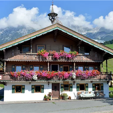 Ein traditionelles bayerisches Haus mit bunten Blumen auf dem Balkon. Im Hintergrund sind grüne Wiesen und Berge zu sehen.