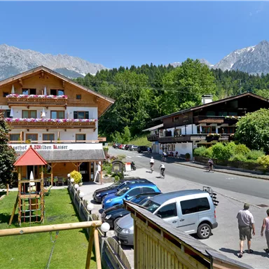 Ein malerisches Gebäude mit Balkon und schönem Garten, umgeben von Bergen. Menschen spazieren entlang der Straße unter blauem Himmel.