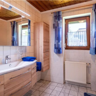 A modern bathroom with wooden shelves and large windows. The walls are light-tiled, and there is a shower as well as a sink.