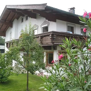 An idyllic house in alpine style with a wooden balcony. The garden is green and blooming, surrounded by trees and plants.