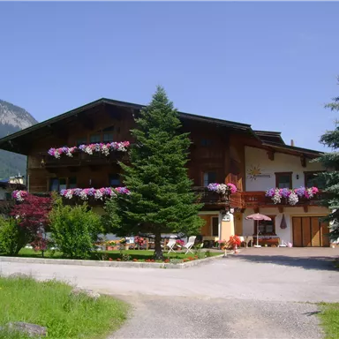 A charming wooden house surrounded by trees and flowers. The clear sky and the mountains in the background create a peaceful atmosphere.
