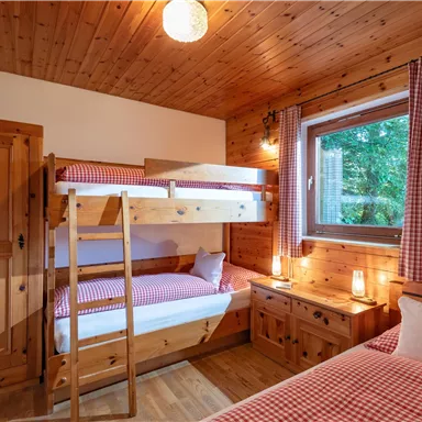 A cozy wooden bedroom with a bunk bed and a single bed. The window offers a view of nature, and the decor feels inviting and warm.