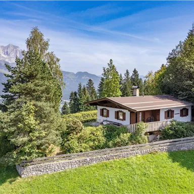 A charming house in the mountains, surrounded by lush greenery and tall trees. In the background, the majestic mountains can be seen and the sky is clear and blue.