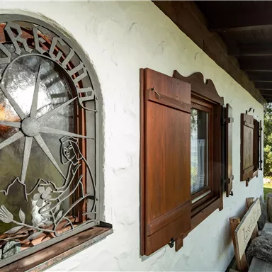 A traditional wooden house with windows and artistic glass design. The surroundings are green and surrounded by trees.