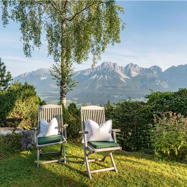 Two lounge chairs are placed on a meadow with a view of majestic mountains in the background. The surroundings are green and invite relaxation.