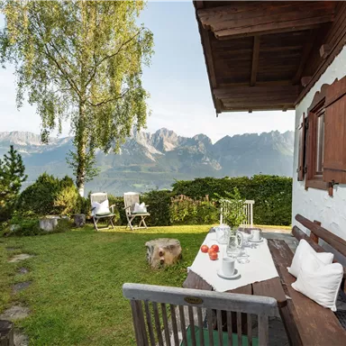 An inviting garden with a view of the mountains. A table is set with glasses and a fruit bowl, surrounded by trees and lawn.