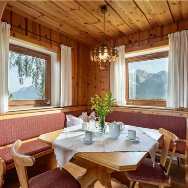 A cozy dining area made of wood with a round table and benches. Through the windows, you can see the mountains and the clear weather.