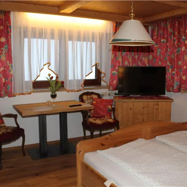 A cozy room with wooden details, a double bed, and a table with two chairs. Colorful curtains hang in front of the windows, and a TV is mounted on the wall.