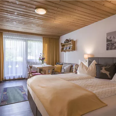 A cozy hotel room with a large double bed and wooden ceiling. There are windows with curtains and a small dining area.