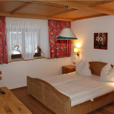 A cozy bedroom with a double bed and wooden furniture. The walls are decorated with colorful curtains, and there is a small seating area.