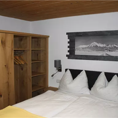 A cozy bedroom with a double bed and two large pillows. The room features a wooden wardrobe and a beautiful mountain landscape on the wall.