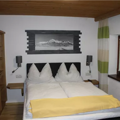 A cozy bedroom with a double bed, wooden cabinets, and modern decoration. The walls are light, and the windows have colored curtains.