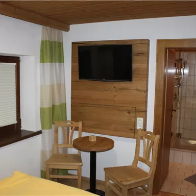 A cozy room with wooden details, two chairs, and a small table. A television hangs on the wall, and there is a bathroom nearby.