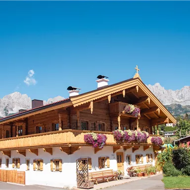 Ein traditionelles, holzverkleidetes Haus mit blühenden Blumen auf dem Balkon. Im Hintergrund sind majestätische Berge und ein klarer Himmel zu sehen.