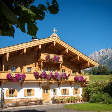 Ein traditionelles Holzhaus mit bunten Blumenampeln und einem klaren blauen Himmel. Im Hintergrund sind beeindruckende Berge zu sehen.