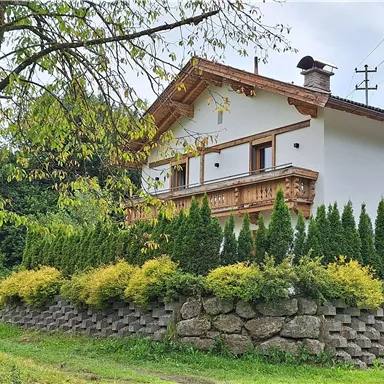 A charming house with a traditional balcony and a well-kept garden. Surrounding it are tall, green plants and a peaceful landscape.