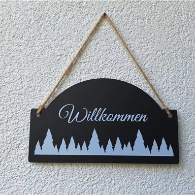 A black sign with the inscription "Welcome" and a stylized mountain landscape of white fir trees below. The sign hangs on a string.