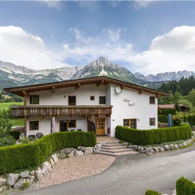 Ein charmantes Haus in den Bergen mit einem gepflegten Garten. Im Hintergrund sind majestätische Berge und ein klarer Himmel zu sehen.