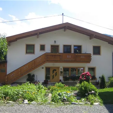 A pretty house with a wooden balcony and plants in the foreground. The surroundings are green and mountainous.