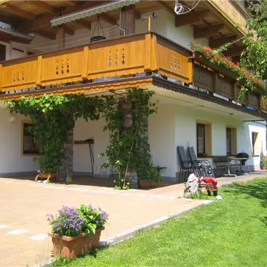 A cozy house with a wooden balcony and green plants. The adjacent garden is equipped with lawn and flowers.