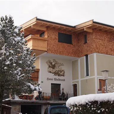 A modern house in alpine style, surrounded by snow-covered trees. The facade is partially clad in wood and features a decorative inscription.