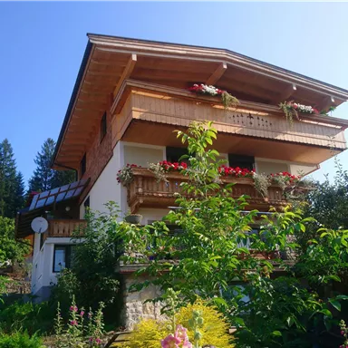 A charming house with several balconies, surrounded by blooming plants and trees. The sky is clear and sunny, enhancing the idyllic atmosphere.