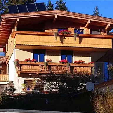 A beautiful wooden house with balconies and flowers. The surroundings are green and offer a clear blue sky in the background.
