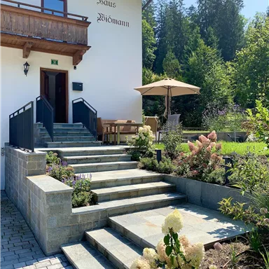 Ein einladender Eingangsbereich mit Steinstufen und grünem Garten. Im Hintergrund steht eine helle Fassade mit einem Balkon.