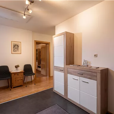 A modern hallway with wooden flooring and minimalist furniture. On display are a chest of drawers, chairs, and a beautiful wall painting.