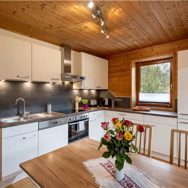 A modern kitchen with a wooden ceiling and light furniture. On the table stands a vase with colorful flowers.