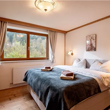 A cozy bedroom with a large bed and a window overlooking nature. It is bright and inviting, with a warm wooden ceiling and soft colors.