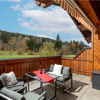 A cozy balcony with modern furniture and a view of a green landscape. The sky is blue with some clouds and the atmosphere is inviting.