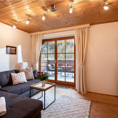 A cozy living room with a gray couch and a table. Large windows with curtains offer a view of a balcony.