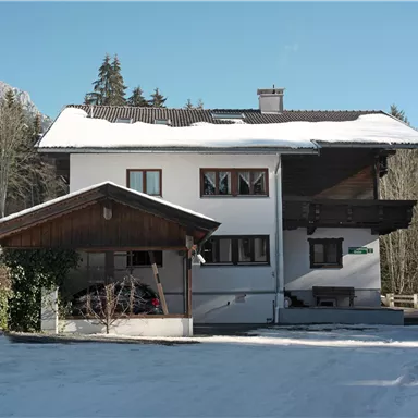 A two-story residential building with a snow-covered roof. In the background, there are snow-covered mountains and a clear blue sky.