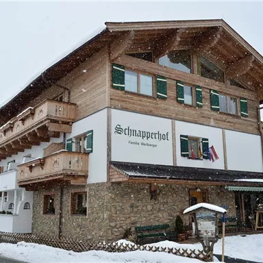 Ein hübsches, zweistöckiges Holzgebäude im Schnee. Es trägt den Namen "Schnapperhof" und hat grüne Fensterläden.