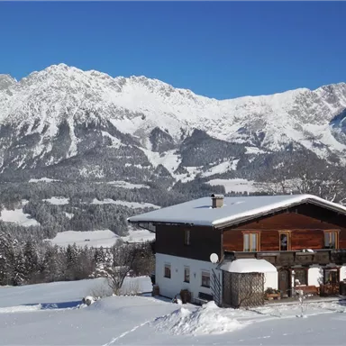 A rustic wooden house in the snow, surrounded by majestic mountains. The sky is clear and blue.