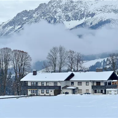 A cozy house in the snow, surrounded by mountains and mist. The landscape is wintry and peaceful.