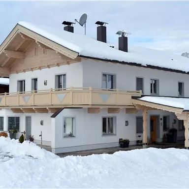 A pretty house with a wooden balcony, surrounded by snow. In the background, there are mountains and a cloudy sky.
