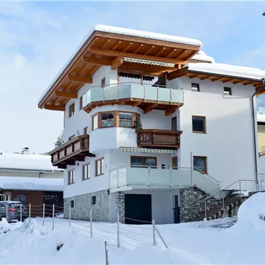 A modern house in the snow, surrounded by other buildings. The balcony and the wooden cladding give the building a charming alpine style.