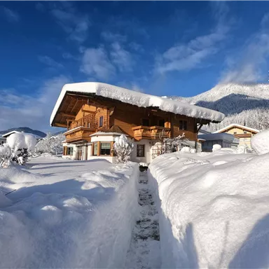 A cozy, two-story wooden house in a snowy landscape. High snow and blue sky surround the idyllic scene.