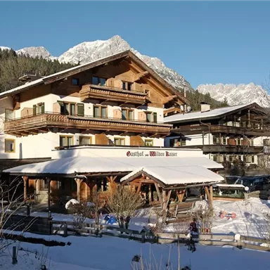 Ein traditionelles österreichisches Hotel im Winter mit schneebedecktem Gelände. Im Hintergrund sind Berge und klare, blaue Himmel zu sehen.