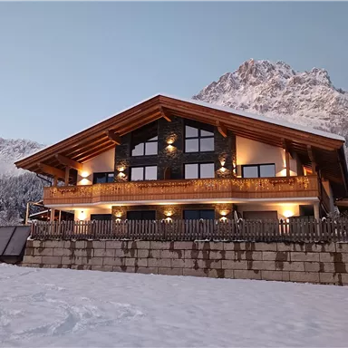 Ein schönes chalet im Schnee, umgeben von schneebedeckten Bergen. Die Fassade ist aus Holz und hat große Fenster, die den Blick nach draußen ermöglichen.