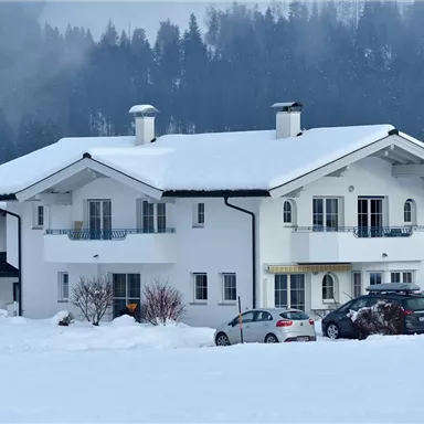 A beautiful house in the snow with a white roof. In the background, snow-covered trees and mountains can be seen.