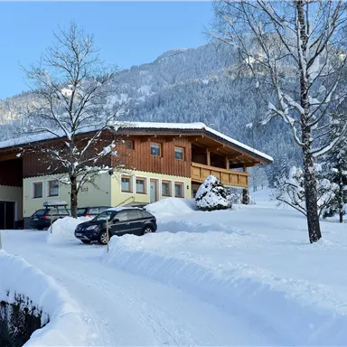 A snow-covered house in the mountains with trees in winter. The path to the house is surrounded by snow.
